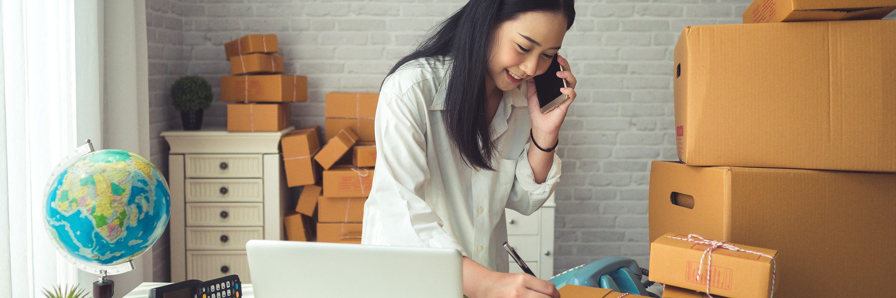 Woman in business casual clothing leaning over a notepad and taking notes inside an office with packages and other office equipment. 